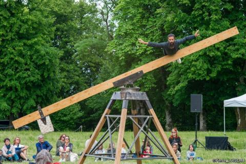 Artiste en équilibre sur une poutre, surplombant du public assis dans l'herbe